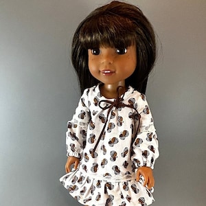 以下が含まれることがあります： 茶色の髪と茶色の目をした人形。茶色の模様が入った白いドレスを着ています。ドレスは長袖で、裾にフリルがあり、首元に茶色のリボンが付いています。人形は灰色の背景の前に立っています。