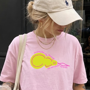 Puede incluir: Una camiseta de manga corta de color rosa claro con un gráfico amarillo y magenta de una bombilla con el texto Finding the way through the smallest spaces, junto con una gorra de béisbol beige con un pequeño logotipo azul marino y un bolso tote beige.