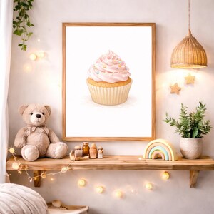 May include: A framed watercolor print of a cupcake with pink frosting and sprinkles. The cupcake is in a light-colored paper cup. The print is displayed on a wooden shelf with a teddy bear, decorative items, and a string of lights.
