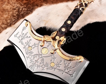 May include: A silver and gold-toned metal hammer with intricate engravings and a leather-wrapped handle. The hammer features a detailed design on the head and a decorative gold accent at the top of the handle. The handle is wrapped in dark brown leather.