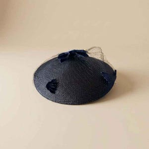 May include: A dark navy blue circular straw hat featuring a low profile and a flat crown. The hat is adorned with small, dark navy blue velvet floral accents and a delicate, fine-mesh netting veil that partially covers the top surface.