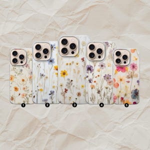 Op de afbeelding: Vijf telefoonhoesjes met bloemmotieven. Elk hoesje heeft een andere opstelling van gedroogde bloemen in verschillende kleuren, waaronder geel, paars, roze en wit, tegen een transparante of gebroken witte achtergrond. De hoesjes zijn genummerd van 1 tot 5.
