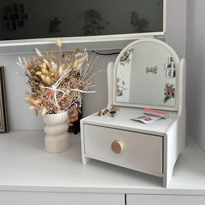 Könnte beinhalten: Ein weißer Holzschminktisch mit einer Schublade und einem runden Holzknauf. Er hat einen bogenförmigen Spiegel und ist mit kleinen Gegenständen dekoriert. Links steht eine Vase mit Trockenblumen.