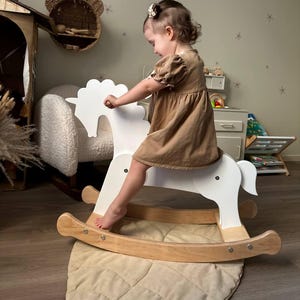 May include: A white wooden rocking horse with a natural wood base. The horse has a simple design and is set on a beige padded mat. The image shows a child wearing a brown dress.
