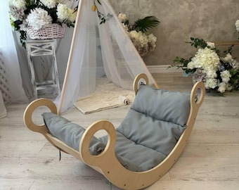 May include: A curved wooden rocker with a light-colored finish and circular cutouts on the sides. The rocker features a soft, padded gray cushion that lines the interior, providing a comfortable seat for resting or gentle rocking motions.