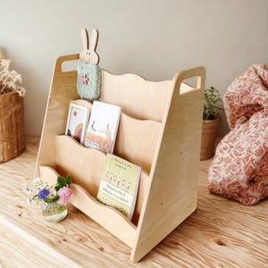 May include: A light-colored wooden bookshelf with three compartments, holding books and a small framed picture. A decorative bunny and a small vase of flowers are also present. The bookshelf has a handle and a wavy top edge.