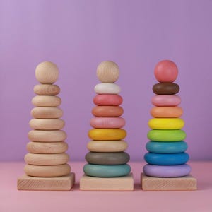 May include: Three wooden stacking toys with colorful rings and a wooden sphere on top. The toys are arranged on a pink surface against a purple background. The rings are in various colors, including natural wood, pink, yellow, green, and blue.