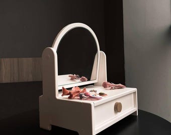May include: A white wooden vanity with a round mirror and a small drawer. The vanity is decorated with small, pink decorative objects. The drawer has a round, wooden knob. The vanity is on a black surface.