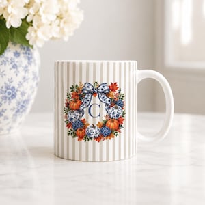 Puede incluir: Una taza de cerámica con un patrón de rayas verticales en beige y blanco. El centro presenta una corona de calabazas naranjas, calabazas con motivos florales azules y blancos, hojas de otoño y bayas, con un lazo azul y blanco y la letra C.