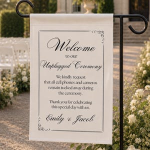 Peut inclure: Un panneau de mariage rectangulaire blanc suspendu à un piquet de jardin en métal noir. Le panneau présente un texte noir indiquant : Welcome to our Unplugged Ceremony. We kindly request that all cell phones and cameras remain tucked away.