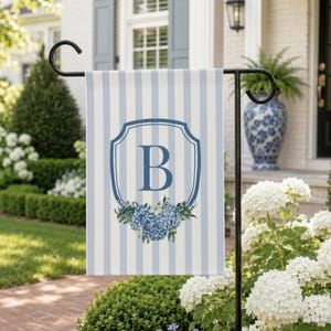 May include: A rectangular garden flag featuring a light blue and white vertical striped pattern. In the center, a dark blue shield-shaped crest frames the letter B, positioned above a cluster of blue hydrangea flowers and green leaves.