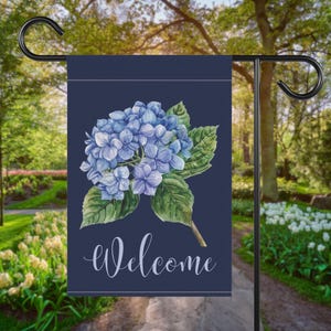 May include: A navy blue garden flag featuring a watercolor illustration of blue hydrangea flowers and green leaves. The word "Welcome" is written in cursive script. The flag is hanging on a black metal stand in a garden setting.