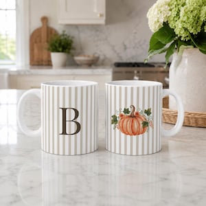 May include: Two white ceramic mugs featuring a vertical beige striped pattern. One mug displays a large, dark brown capital letter B, while the other mug features a detailed illustration of an orange pumpkin surrounded by green leaves and small berries.