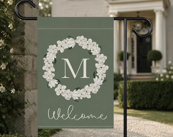 Könnte beinhalten: Eine rechteckige Gartenflagge in Salbeigrün mit einem zentralen weißen Blumenkranz, der den Großbuchstaben M in einer Serifenschrift umschließt. Unter dem Kranz steht das Wort Welcome in weißer Schreibschrift. Die Flagge hängt an einem schwarzen Ständer.