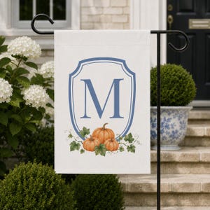 May include: A white rectangular garden flag featuring a blue outlined crest with a large blue letter M in the center. Below the crest are three orange pumpkins with green leaves and vines, displayed on a black metal garden flag stand.
