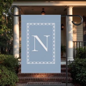 May include: A light blue garden flag with a white decorative border and a large white letter "N" in the center. The flag is hanging on a black metal stand in front of a house.