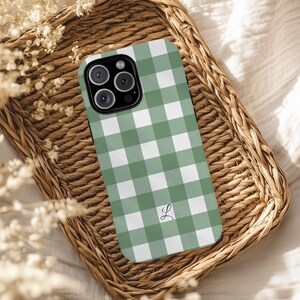 May include: A green and white gingham pattern phone case with a black camera module. The case features a cursive letter "L" in the lower right corner. The phone case is resting on a woven wicker tray.