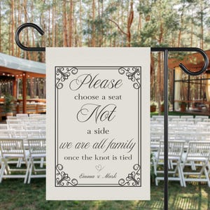May include: A cream wedding garden flag with black text. The flag reads "Please choose a seat, Not a side, we are all family once the knot is tied." The flag is framed with decorative flourishes and includes the names "Emma & Mark".
