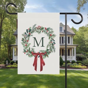 May include: A cream garden flag displays a Christmas wreath design with green leaves, red berries, and a burgundy bow. The letter "M" is in the centre of the wreath. The flag is on a black metal stand in a garden.