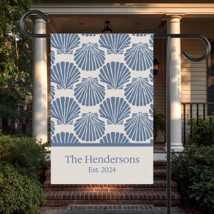 May include: A garden flag featuring a repeating pattern of blue seashell designs on a cream background. The bottom section displays the text "The Hendersons" and "Est. 2024". The flag is displayed on a black metal stand.