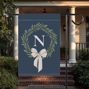 May include: A navy blue garden flag featuring a wreath of green leaves around the letter "N". A white bow with tan stripes is below the letter. The flag is displayed on a black metal stand in front of a house.