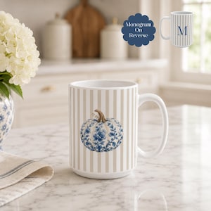 Puede incluir: Una taza de cerámica blanca con rayas verticales beige y una ilustración central de una calabaza con motivos florales azules y blancos. Un recuadro muestra el reverso con un monograma azul de la letra M. El texto Monogram On Reverse es visible.