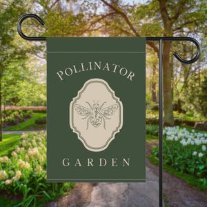 Puede incluir: Una bandera de jardín verde oscuro con las palabras "POLLINATOR GARDEN" en blanco. Una ilustración de abeja beige está centrada en la bandera. La bandera está colgada de un poste de jardín de metal negro.