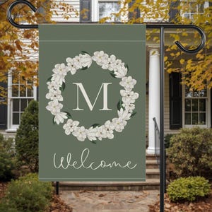 Puede incluir: Una bandera de jardín verde con un diseño de corona floral blanca alrededor de la letra "M". La palabra "Welcome" está escrita en cursiva debajo de la corona. La bandera se muestra en un soporte de metal negro.