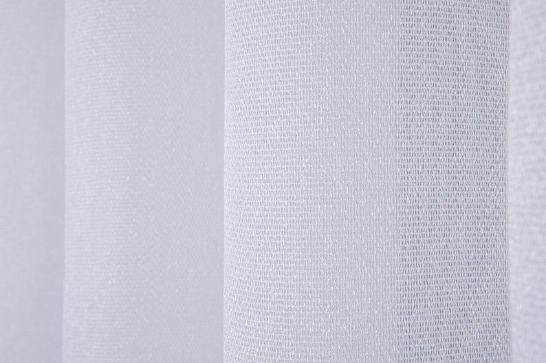 Pode incluir: Um tecido de cortina branco com uma textura de malha fina e semitransparente com dobras verticais. O material exibe um padr&atilde;o de tecelagem leve e consistente que cria sombras e reflexos sutis na superf&iacute;cie do t&ecirc;xtil pendurado.