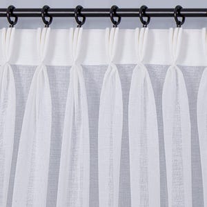 Könnte beinhalten: Weiße, transparente Vorhänge mit einem plissierten Design hängen an einer schwarzen Metallstange mit schwarzen Ringen. Die Vorhänge bestehen aus einem leichten Stoff, der das Durchscheinen von Licht ermöglicht. Thema: Heimdekoration.