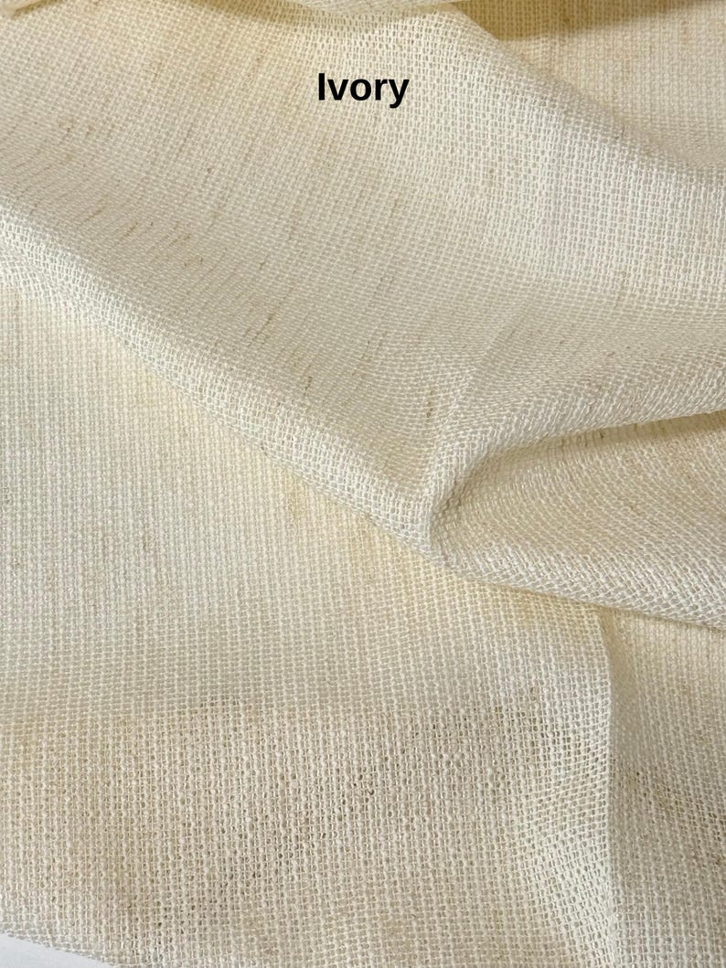 Pode incluir: Um tecido de cor marfim com uma textura vis&iacute;vel, grossa e de trama aberta. O material est&aacute; drapeado em dobras suaves, criando sombras e reflexos naturais na superf&iacute;cie. A palavra "Ivory" est&aacute; impressa em texto preto sobre o tecido.