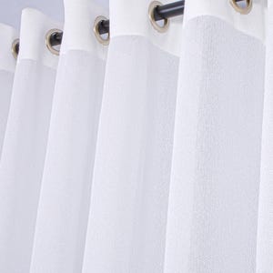 Pode incluir: Um conjunto de cortinas brancas em tecido texturizado e semitransparente com pregas verticais. O topo &eacute; refor&ccedil;ado com uma faixa de tecido branco s&oacute;lido e inclui ilh&oacute;s met&aacute;licos circulares espa&ccedil;ados num var&atilde;o de cortina escuro.