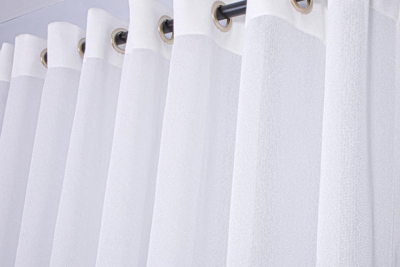 Pode incluir: Um conjunto de cortinas brancas em tecido texturizado e semitransparente com pregas verticais. O topo &eacute; refor&ccedil;ado com uma faixa de tecido branco s&oacute;lido e inclui ilh&oacute;s met&aacute;licos circulares espa&ccedil;ados num var&atilde;o de cortina escuro.