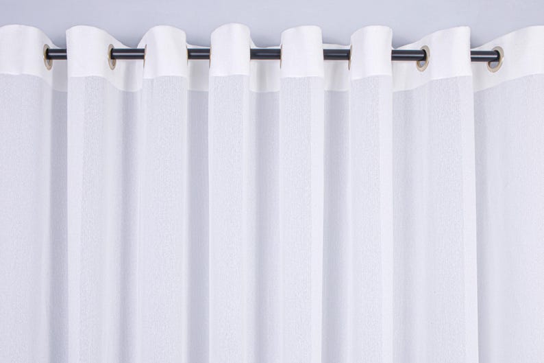 Pode incluir: Um painel de cortina branco com uma faixa superior branca s&oacute;lida e ilh&oacute;s met&aacute;licos espa&ccedil;ados uniformemente. A cortina est&aacute; pendurada numa vara horizontal escura e fina, criando pregas verticais e uma apar&ecirc;ncia texturizada semitransparente.