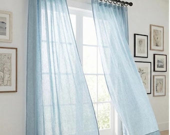 Pode incluir: Um par de cortinas azul-claras semitransparentes penduradas numa vara de metal escuro com argolas circulares. O tecido tem uma trama texturizada subtil que permite a passagem da luz, criando um aspeto suave e arejado na janela.