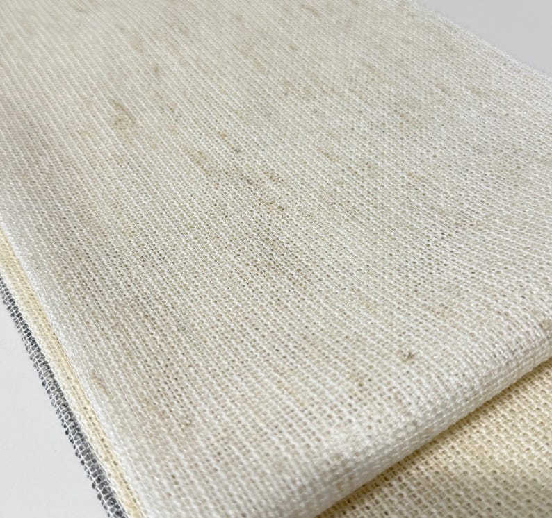 Pode incluir: Uma pe&ccedil;a de tecido bege claro dobrada com uma textura tecida vis&iacute;vel. O material apresenta uma apar&ecirc;ncia natural e ligeiramente irregular com subtis manchas castanhas em toda a trama, criando uma superf&iacute;cie consistente de tons neutros.