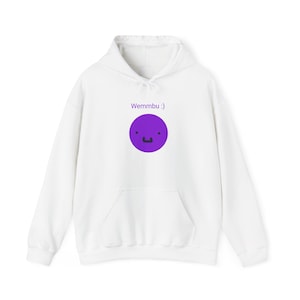 Puede incluir: Una sudadera con capucha blanca que presenta un gráfico de círculo morado en el pecho. El gráfico muestra una cara sencilla con dos puntos pequeños para los ojos y una boca negra pequeña. Sobre el círculo morado, el texto Wemmbu :) está impreso.