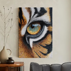 Puede incluir: Primer plano de una impresión en lienzo que muestra el ojo de un tigre. El ojo es una vibrante mezcla de amarillo, azul y marrón, con pelaje detallado y rayas blancas y negras. La obra de arte se exhibe en una pared.