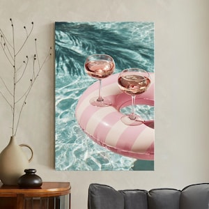 May include: Two stemmed wine glasses filled with rosé wine rest on a pink and white striped inflatable ring in a swimming pool. The water is a clear blue, and the scene is lit by sunlight.