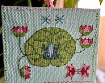 May include: A cross-stitch embroidery featuring a green lily pad with a blue frog on top, surrounded by pink water lilies and two blue dragonflies. The embroidery is framed with a light blue background and a pink border.