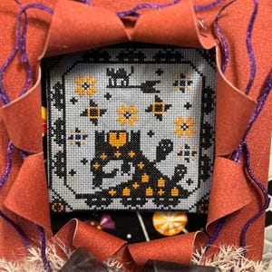 Puede incluir: Un punto de cruz enmarcado con un gato negro con manchas naranjas, estrellas y calabazas sobre un fondo azul claro. El marco está rodeado de papel naranja con detalles de purpurina morada y un lazo negro, creando un tema de Halloween.