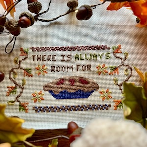 Puede incluir: Obra de arte de punto de cruz con la frase "THERE IS ALWAYS ROOM FOR" sobre una ilustración de tarta. La tarta tiene una corteza marrón y una base azul, con relleno rojo. La obra está enmarcada por follaje de otoño y bellotas.
