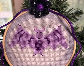 May include: A cross-stitch ornament featuring a purple bat with outstretched wings, set against a light purple background. The design is framed in a wooden hoop and adorned with a purple glitter ribbon and a cluster of black and purple decorative beads. The ornament is displayed on a green, leafy background.