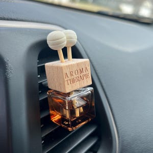 Könnte beinhalten: Ein brauner Glas-Lufterfrischer für das Auto mit einem Holzdeckel und zwei weißen Diffusionskugeln. Der Holzdeckel trägt die Gravur "Aroma Therapie".