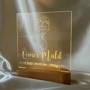 May include: A personalized night light with a baby footprint design. The clear acrylic panel is illuminated by a warm glow, and features the name "Omer Malik", date, time, weight (3560 grams), and height (55 cm). The base is made of wood.