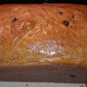 Op de afbeelding: Een bruin brood met een glanzende korst. Het brood heeft een rechthoekige vorm en lijkt te zijn bezaaid met donkere rozijnen. Het brood ligt op een lichtgekleurd oppervlak.