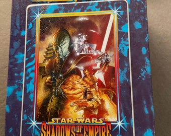 Puede incluir: Una caja de metal azul y morado con el texto "Star Wars Shadows of the Empire" y "Embossed Metal Collector Cards". La caja presenta una ilustración colorida de personajes del universo Star Wars.