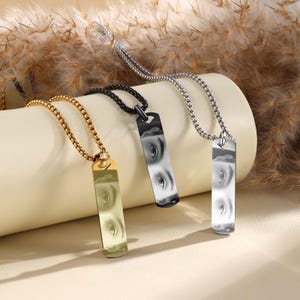 Puede incluir: Tres collares con colgantes rectangulares con un dise&ntilde;o de ojo. Los collares son dorados, negros y plateados, cada uno con una cadena a juego. Los colgantes est&aacute;n orientados verticalmente y tienen una superficie reflectante.