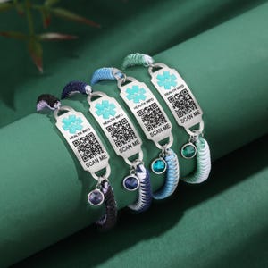 May include: Four medical alert bracelets with woven bands in various colors. Each bracelet features a silver tag with a medical symbol, the words "HEALTH INFO" and "SCAN ME", and a QR code. Each bracelet has a dangling charm.