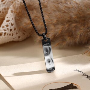 Puede incluir: Un collar con colgante rectangular negro que muestra una imagen en blanco y negro de ojos. El colgante cuelga de una cadena negra. El fondo presenta tonos beige y marr&oacute;n.